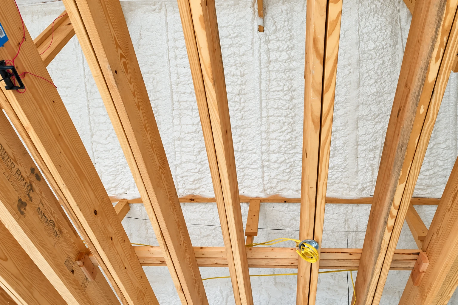 Close-up of white spray foam insulation between exposed wooden roof framing, with electrical wiring and a junction box.