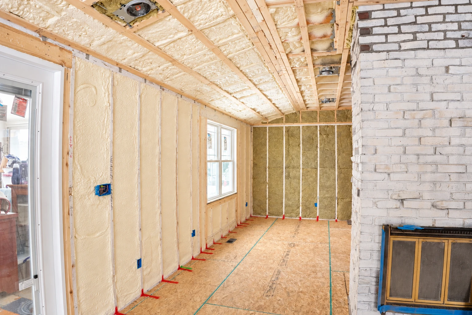 Unfinished room with cream spray foam insulation across walls and ceiling joists, beside a white brick fireplace.