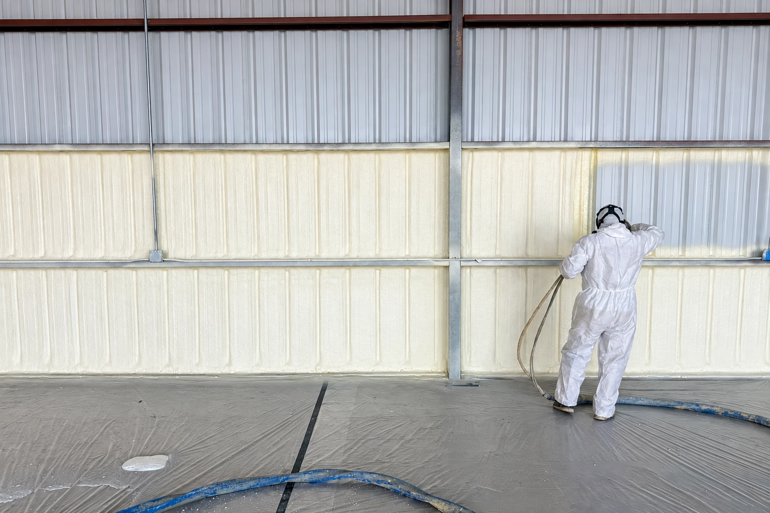 A worker in white protective gear sprays pale foam insulation onto the corrugated metal wall of a large building.