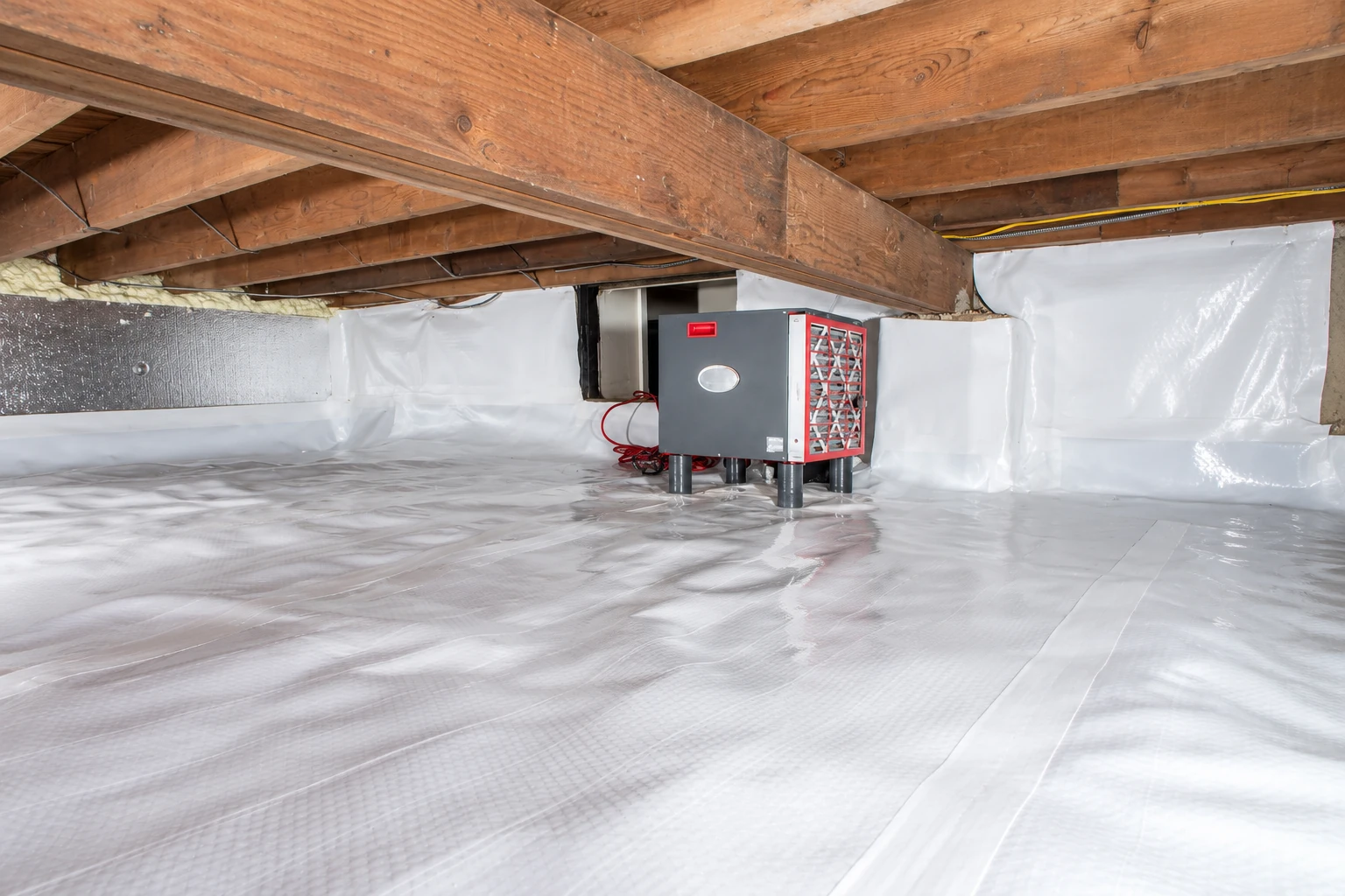 Crawl space with a white plastic vapor barrier covering the floor and walls, wooden joists overhead, and a compact HVAC unit.