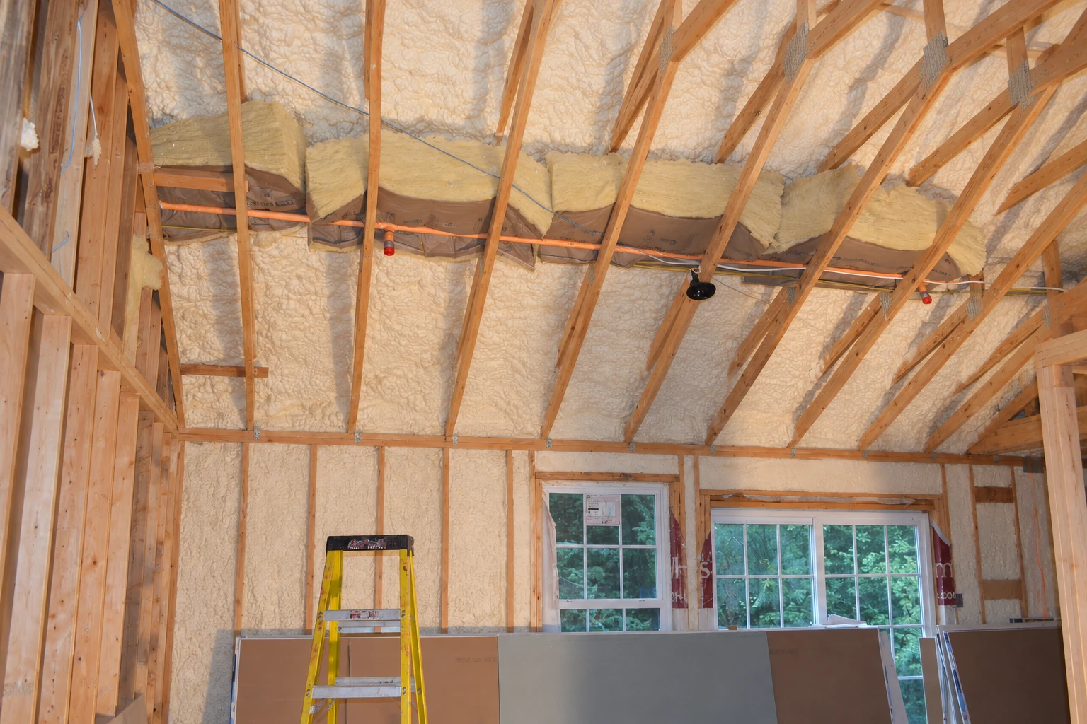 Foam insulation coats the walls and roof framing of an unfinished home, with fiberglass batts resting across the rafters.