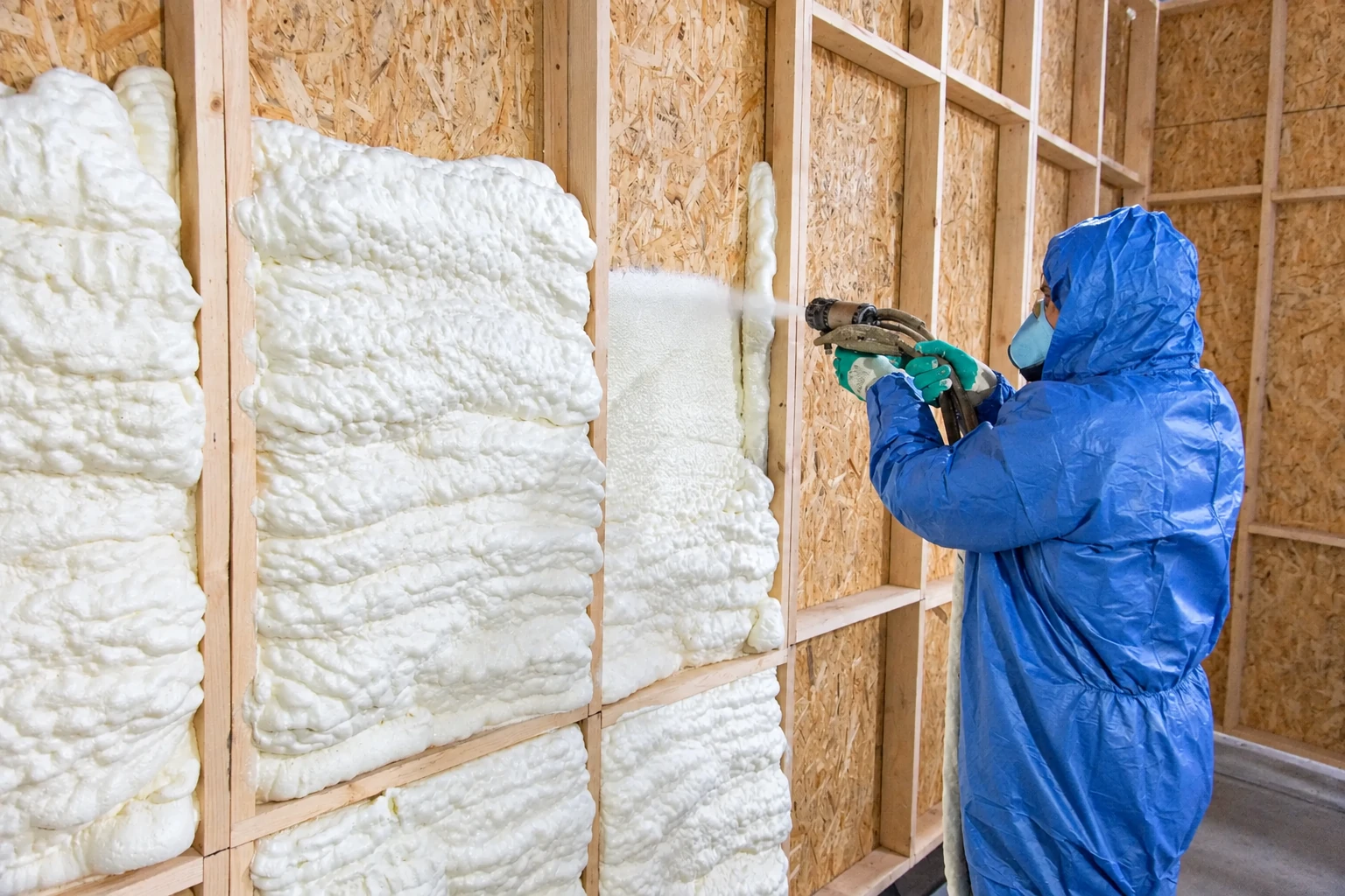 Worker in blue protective gear sprays white open-cell foam insulation into wooden wall cavities.